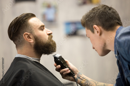 Profile view portrait of handsome young bearded caucasian man getting beard grooming in modern barbershop. Hairdresser with tattoo "one love" serving client, making beard haircut using shaver
