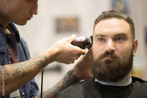 Close-up portrait of handsome young bearded man getting trendy haircut in modern barbershop. Cool male hairstylist with tattoo "born barber" serving client. Indoors shot