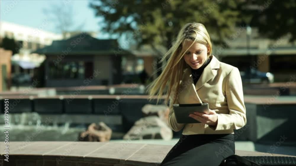 A pretty blond businesswoman sitting by of a city fountain using her digital or electronic tablet smiles at what she sees. A pretty blond businesswoman sitting by of a city fountain using her digital or electronic tablet smiles at what she sees.
