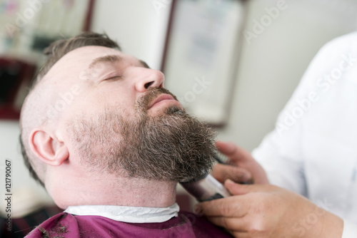 Male barber cuts the beard using clipper of a adult man with a mohawk