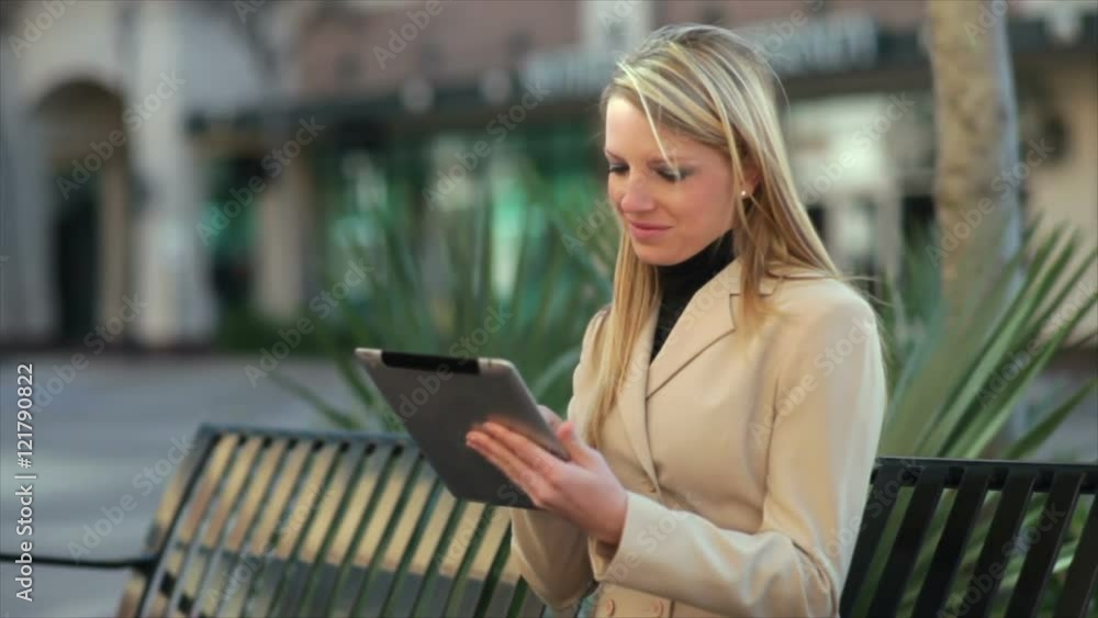 Sitting on a municipal park bench using an electronic tablet a pretty blond businesswoman enjoys the sunshine of a breezy fall day. Sitting on a municipal park bench using an electronic tablet a pretty blond businesswoman enjoys the sunshine of a breezy fall day.
