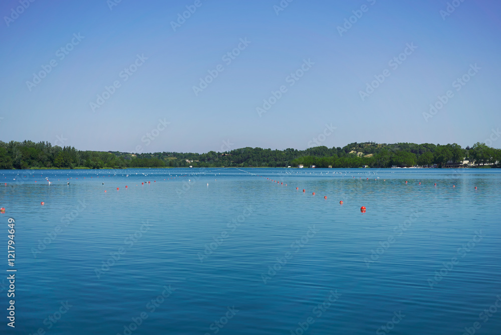 Rowing lane marked by orange and white buoys on a peaceful lake surrounded by hills under a clear blue sky