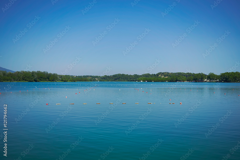 Lines of buoys marking rowing lanes on a peaceful greenish blue lake under a quiet sunny sky