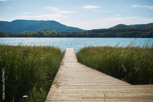 Long wooden pier leads through tall grass to a sparkling blue lake surrounded by wooded hills on a clear summer day