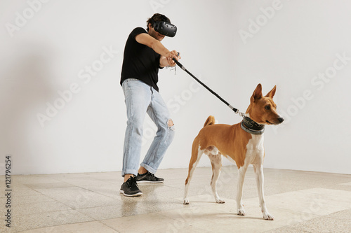 A young gamer in VR headset and black unlabeled t-shirt pulling a leash on a brown and white basenji dog against a white wall background.
