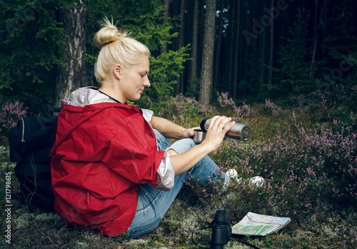 Young active girl tourist drinking tea from a thermos and sitting in the forest. Healthy active lifestyle concept