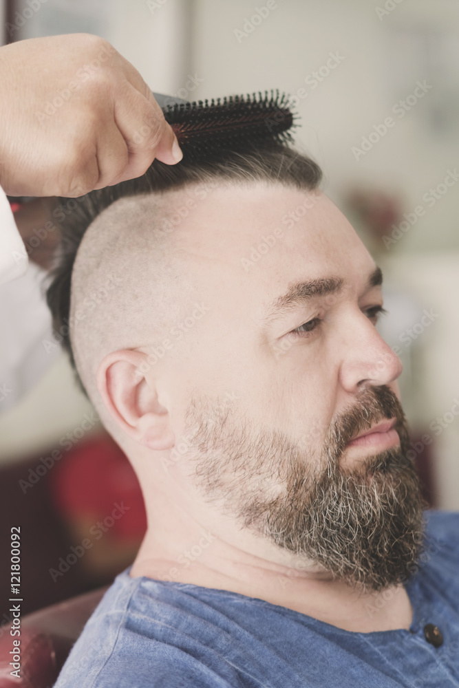 Obraz premium Male barber makes hair styling using a hair dryer of a adult bearded man with a mohawk