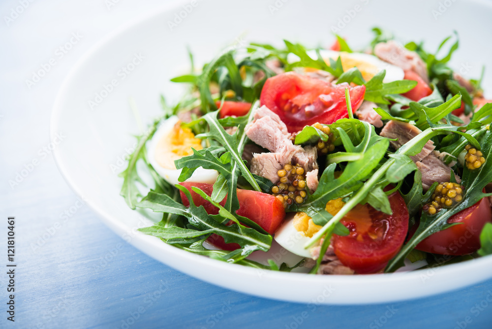 Fresh salad with tuna, tomatoes, eggs, arugula and mustard on blue wooden background close up. Healthy food.