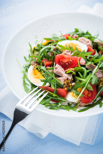 Fresh salad with tuna, tomatoes, eggs, arugula and mustard on blue wooden background close up. Healthy food.