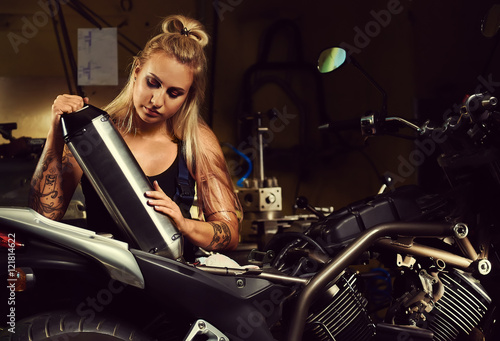 Blond woman mechanic holding a muffler in a motorcycle workshop
