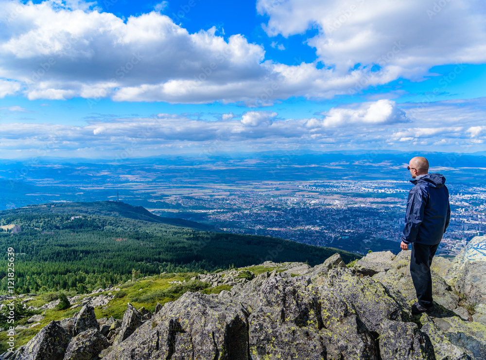 Middle aged man in sports clothing and sunglasses on top of a mountain, overlooking the city of Sofia, Bulgaria from high altitude - beautiful landscape with city shapes in the far background