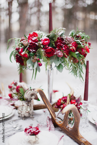 Wedding table setting in marsala colors with plates, cutlery, red floral compositions, candles, velvet napkins, deer horns on table covered with snow