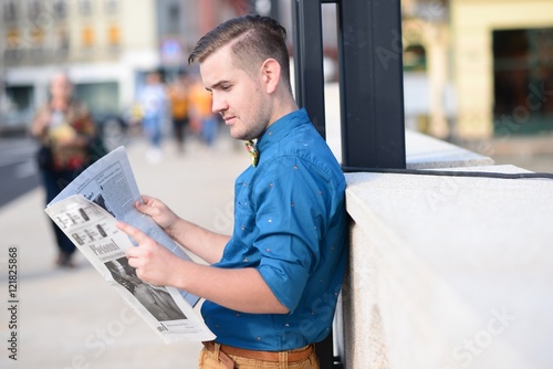Young man reading the newspaper while waiting in a crowded street
