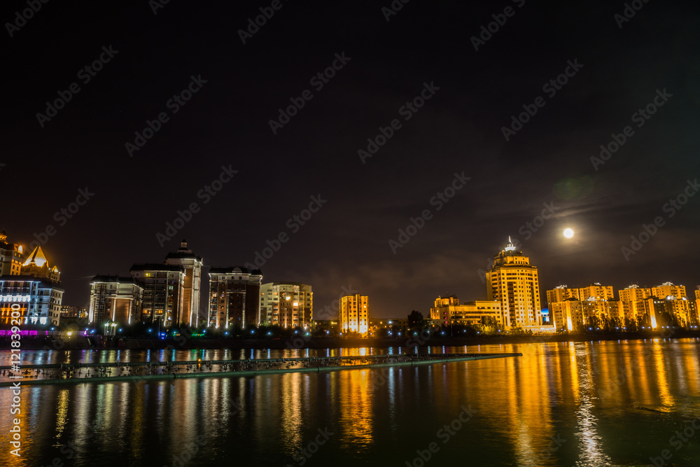 Astana, Kazakhstan. Ishim river embankment in the moon night with buildings and reflection in the water