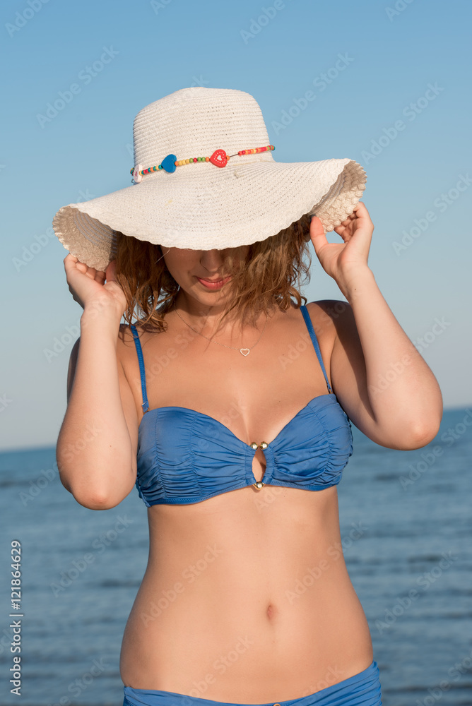 Blond woman wear blue bikini and white hat, clear sky and sea as background