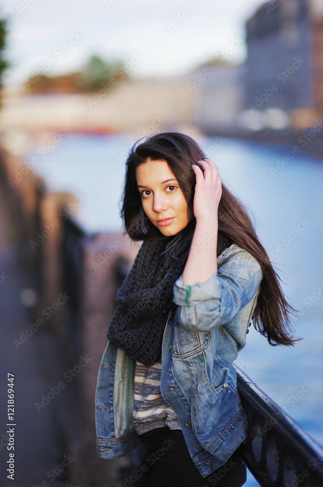 Young beautiful long-haired girl with  brown eyes on the waterfront in the setting sun  a blurred background of the river and the city's architecture.