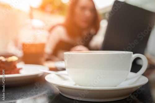 Soft focus morning coffee cup on desk with blur young women working with laptop computer