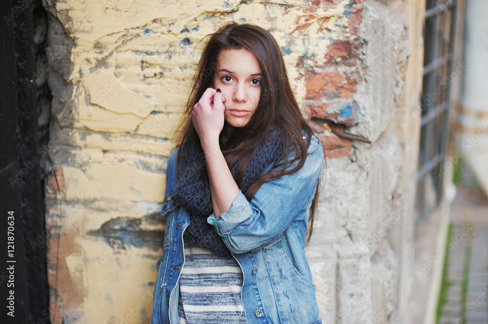 Sad young brown-eyed girl with long hair in a black scarf, and denim jacket against the background of an old brick wall, close-up. Sad young brown-eyed girl with long hair in a black scarf, and denim jacket against the background of an old brick wall, close-up.