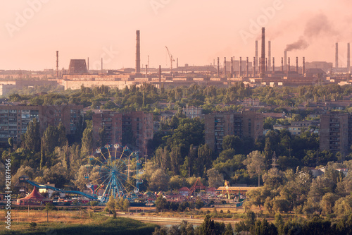 Amusement park with Ferris wheel and buildings on the background of metallurgy plant with smog at sunset. Ecology problems, atmospheric pollutants. Pipes with smoke. Heavy industry factory. Steel mill photo