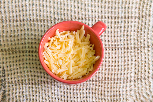 Grated Gouda cheese in red cup on linen tablecloth. Aerial view