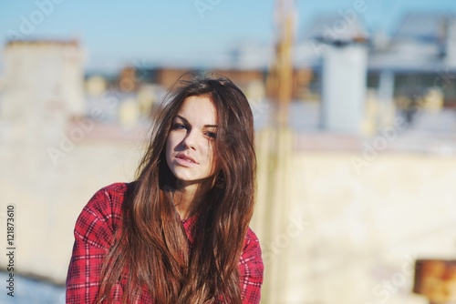 Beautiful brunette girl with long shiny hair blue eyes in a red plaid shirt on the background of the city roofs.