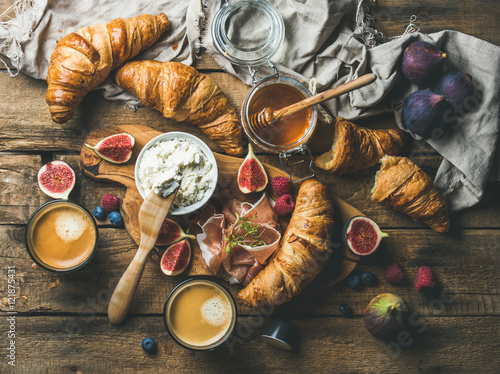 Breakfast with freshly baked croissants, ricotta cheese, figs, fresh berries, prosciutto, honey in glass jar and coffee espresso over rustic wooden background, top view, horizontal composition
