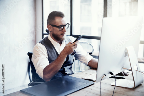 Bearded Stylish Young Man Wearing Glasses White Shirt Waistcoat Working Modern Loft Startup Process.Creative Person Using Smartphone Texting Message.Drawing Tablet Desktop Computer Wood Table.Blurred.