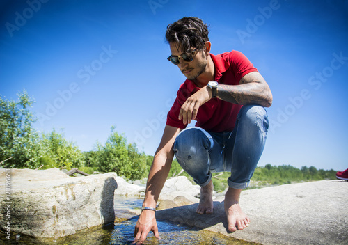 Attractive young man outdoor at river or water stream, sitting on big rock, looking away, with stones in background photo