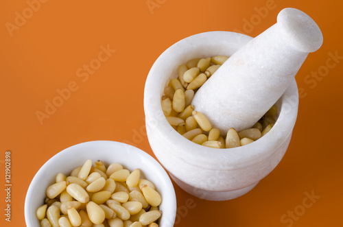 Shelled pine nuts in white bowl and mortar on orange background. Edible seeds of Korean pines family Pinaceae, Pinus koraiensis, in a porcelain bowl and in a marble mortar. Macro food photo close up. photo