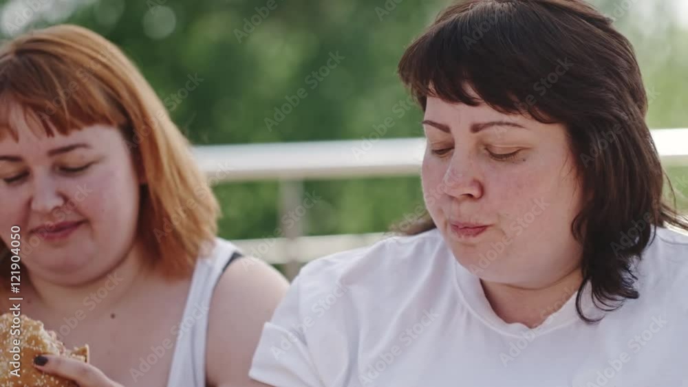 Tracking shot of three fatty women eating burgers and fries and drinking coke from plastic bottle Tracking shot of three fatty women eating burgers and fries and drinking coke from plastic bottle