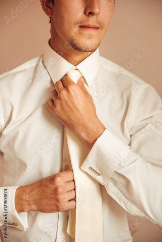 A picture of a groom fixing a fine tie carefully, neat white shirt on. Man corrects belt, fees groom, man's hands, dressing, man buttons pants