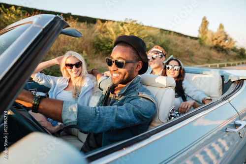 Fototapeta Cheerful young friends driving car and smiling in summer