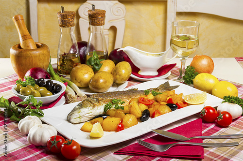 Fried seabass with side dish on a plate, served on a table next to fresh vegetables, olives, olive oil, cutlery and a glass of white wine.