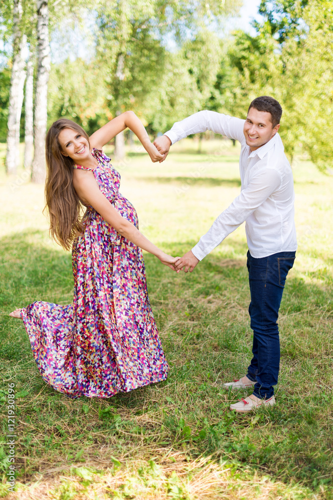 Beautiful pregnant couple happy together expecting a child. Man and woman walking in the park showing heart with hands. Share the love and family, love concept.