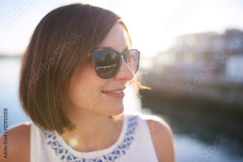 Happy caucasian woman looking off camera, wearing sunglasses and smiling with warm summer light coming through from behind her.