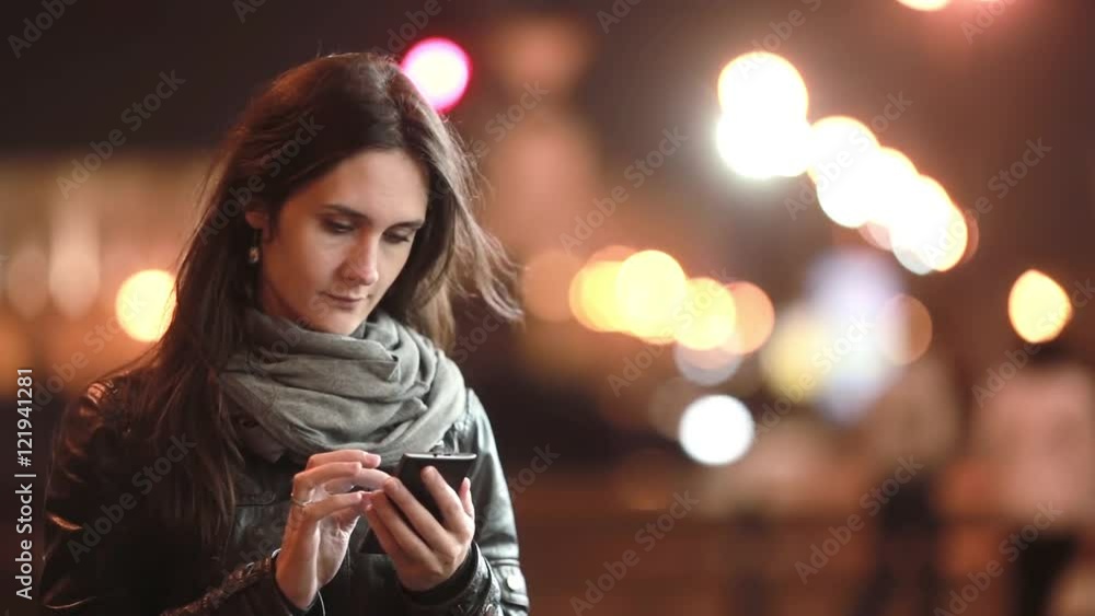 Pretty young woman smiling, using her smartphone at night. Lady in the street in cold weather. Modern technology. Pretty young woman smiling, using her smartphone at night. Lady in the street in cold weather. Modern technology.