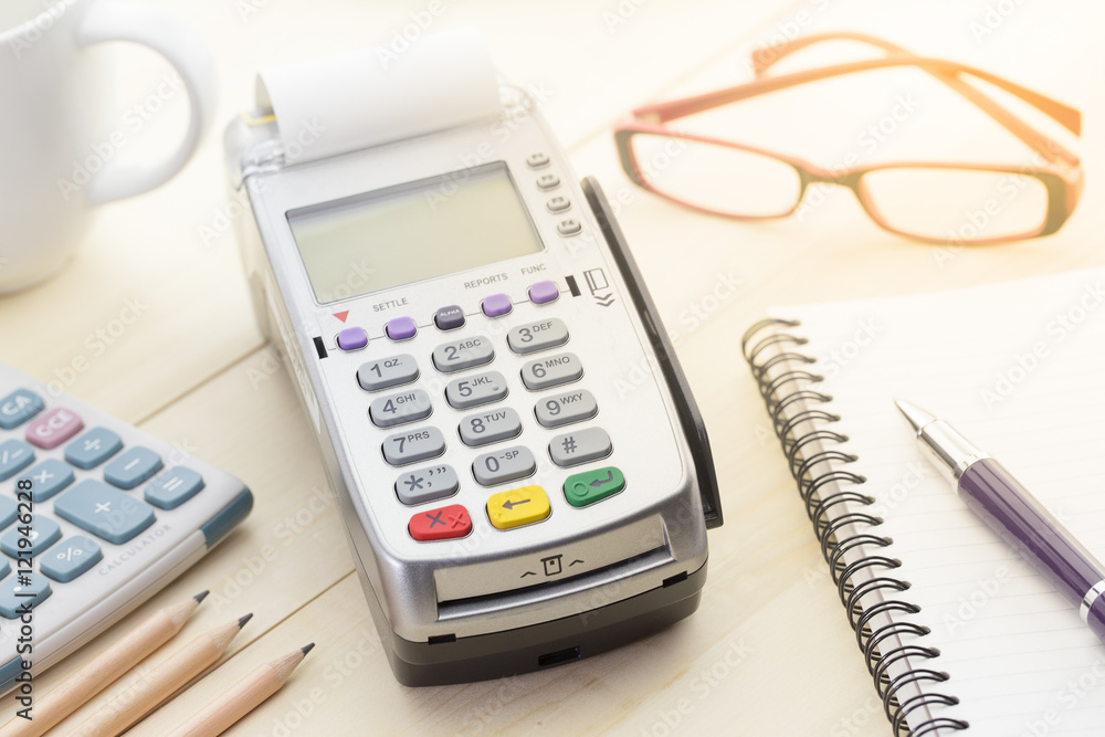 Credit Card Terminal or EDC on cashier wooden table in the store with credit card, pen, calculator and glasses on background. Credit Card Terminal or EDC on cashier wooden table in the store with credit card, pen, calculator and glasses on background.
