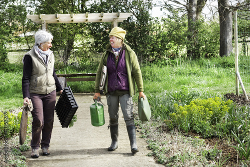 Two women walking along a path carrying kneeler pads and watering can and plant trays. photo