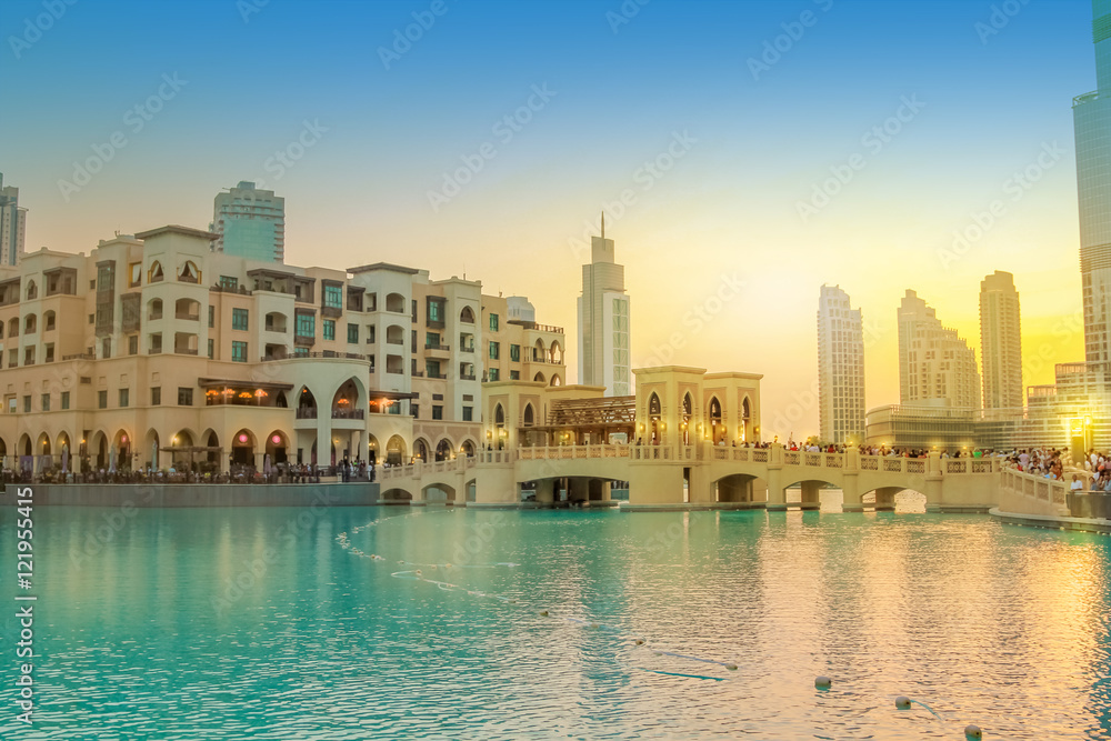 Fototapeta premium Scenic view of Burj Khalifa Lake, a pool where they dance the Dubai Fountain. On background, the bridge that connects the Dubai Mall to Souk Al Bahar. People crowd around the shopping area at sunset.