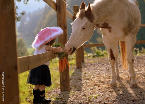 Fototapeta Naklejka Na Ścianę i Meble -  Freunde fürs Leben, kleines süßes Cowgirl mit pinkem Hut füttert Westernpferd mit Möhren