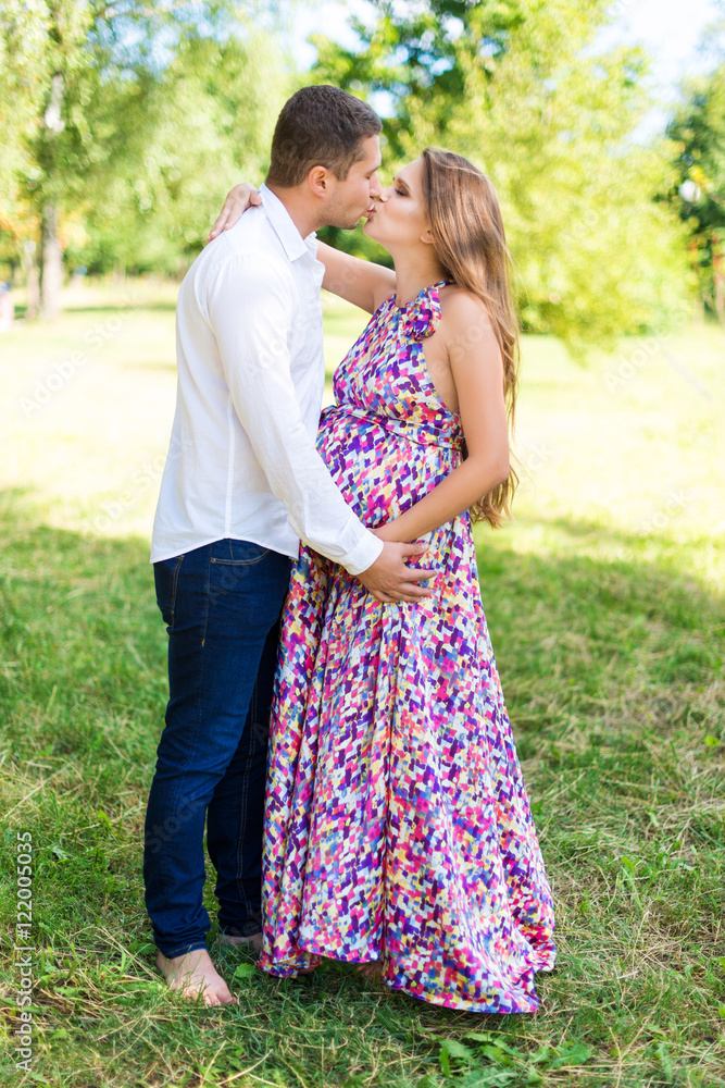 Beautiful pregnant couple happy together expecting a child. Man and woman kissing while walking in the park. Share the love, family, love concept.
