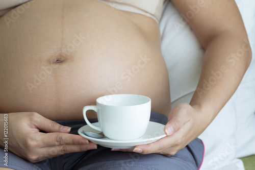 Pregnant woman hands holding cup coffee on belly in bedroom.