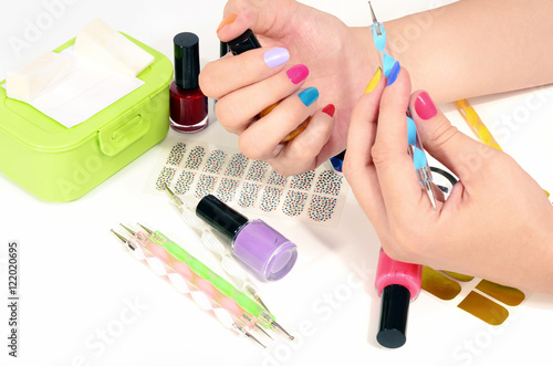 Woman hands doing rainbow manicure.Table full of manicure ustensils,coloured nail polish,nail decals stickers,striping tape,nail file,dotting pens,vinyl sheets.Nail art accesories isolated on white. 