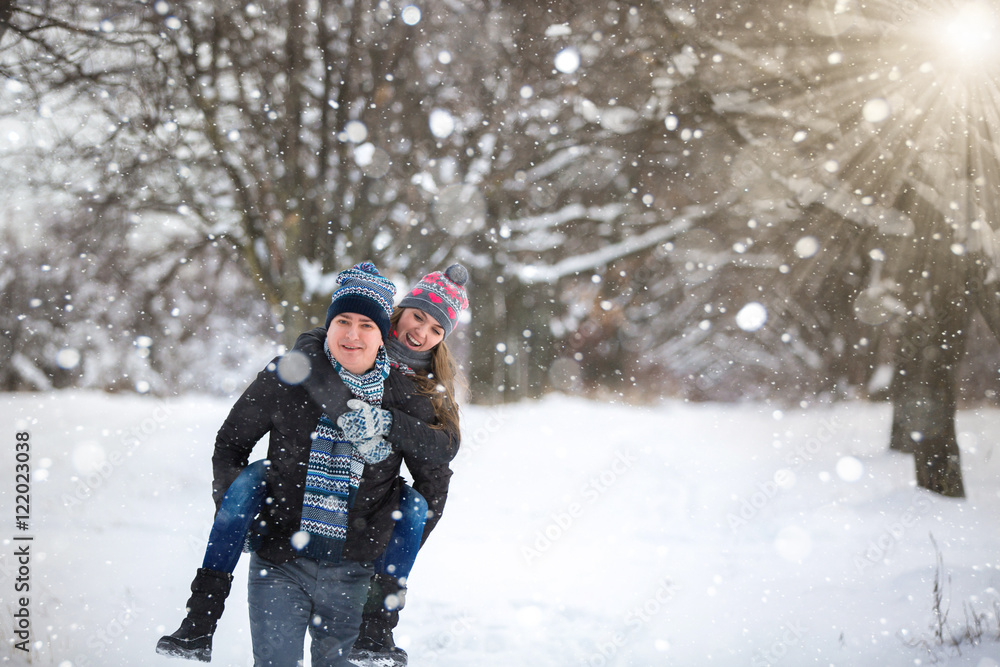 A loving couple is playing in the winter park. A girl sits on the shoulders of men, he flees. Winter snowfall. Valentine's Day, Christmas vacation. Copy space.