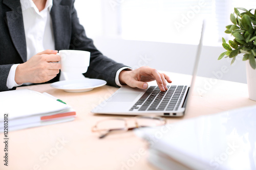 Close up of business woman hands with a cup of coffee is sitting at the table and typing on a laptop computer in the white colored office .