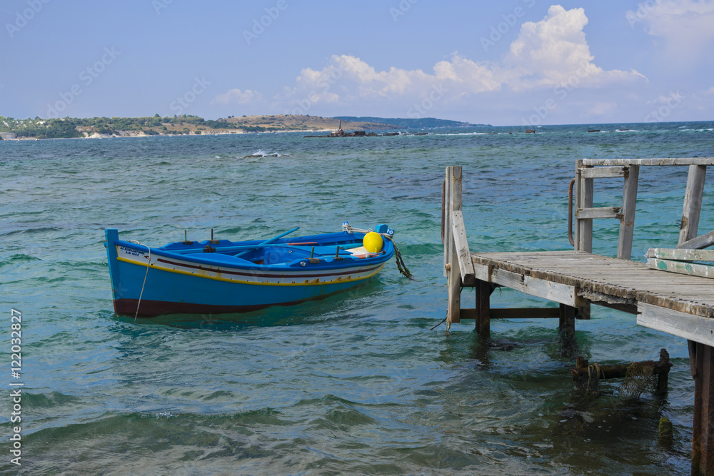 Blue sea fishing boat (vessel) in the old harbor with wooden mooring and island at the background Blue sea fishing boat (vessel) in the old harbor with wooden mooring and island at the background