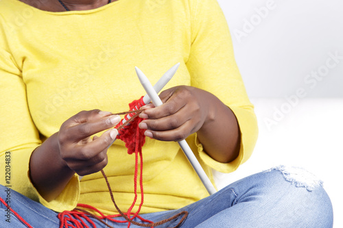 African woman knitting sitting on a white couch. Knitting something with red colour  adding brown.