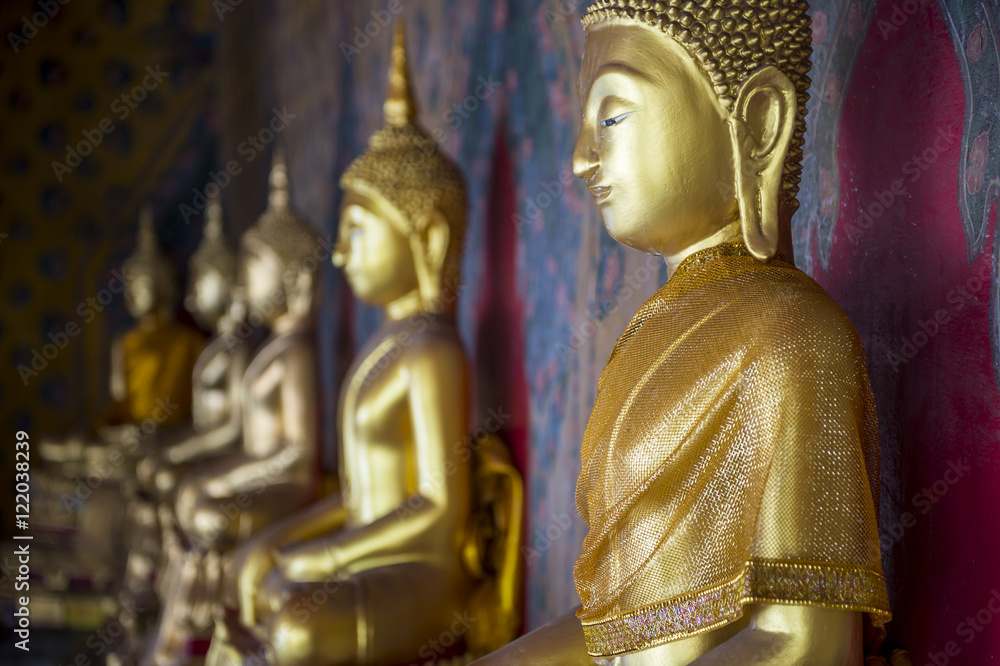 Row of golden seated buddhas wearing yellow sash in front of decorative wall in a Buddhist temple in Bangkok Thailand