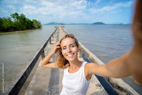 Vacation and technology. Young smiling woman taking selfie while walking on sea bay pier.