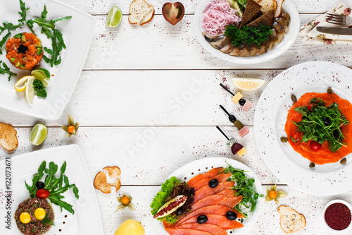 Delicious mediterranean snacks on white table, free space, flat lay. Tasty plates with various salads, smoked fish and meat on white wooden background, copy space for text